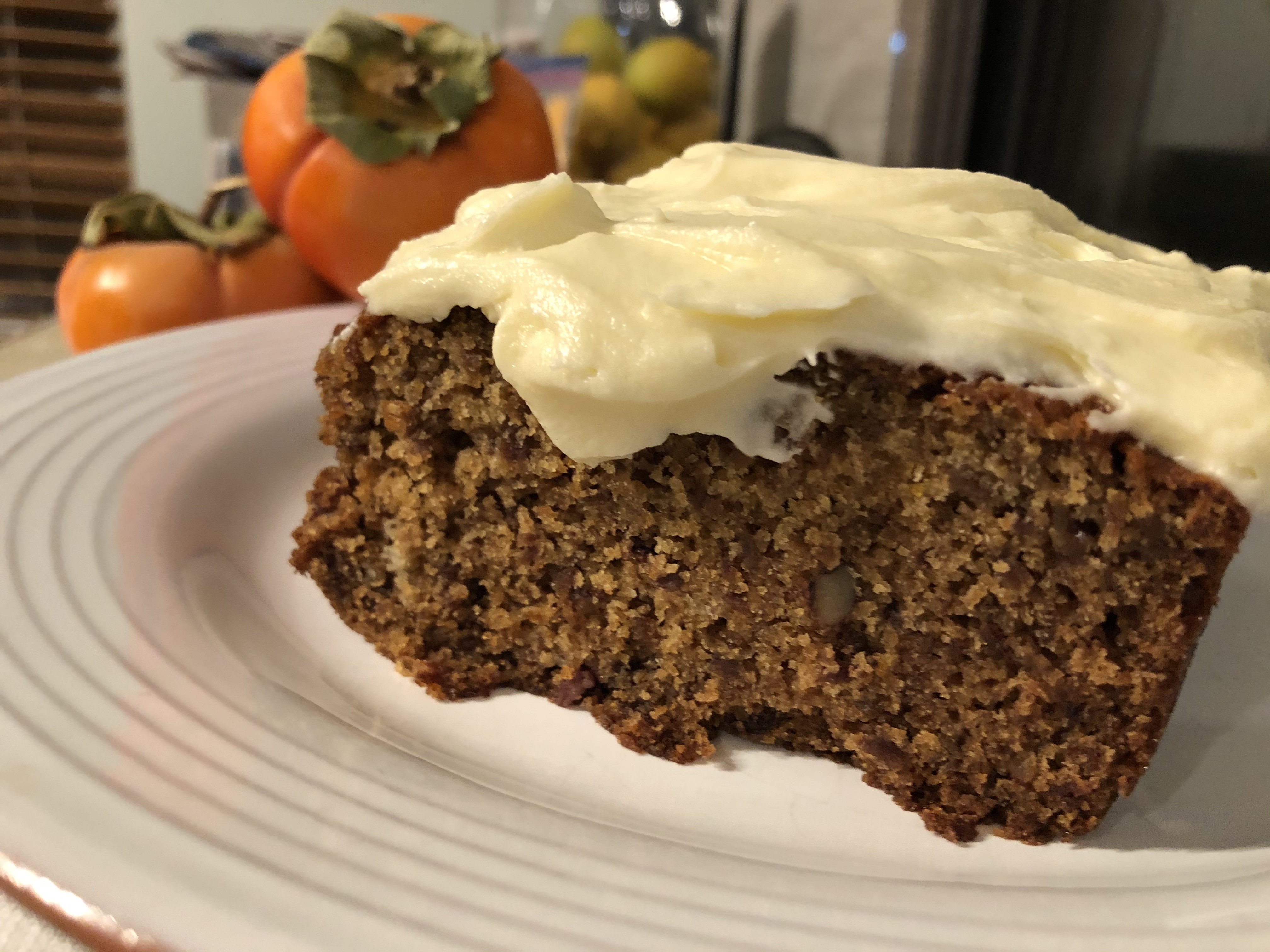 A slice of Fuyu Persimmon Cake with cream cheese frosting, with fresh Fuyu persimmons in the background